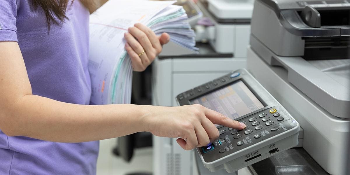 Person operating a multifunction office printer while holding a stack of documents