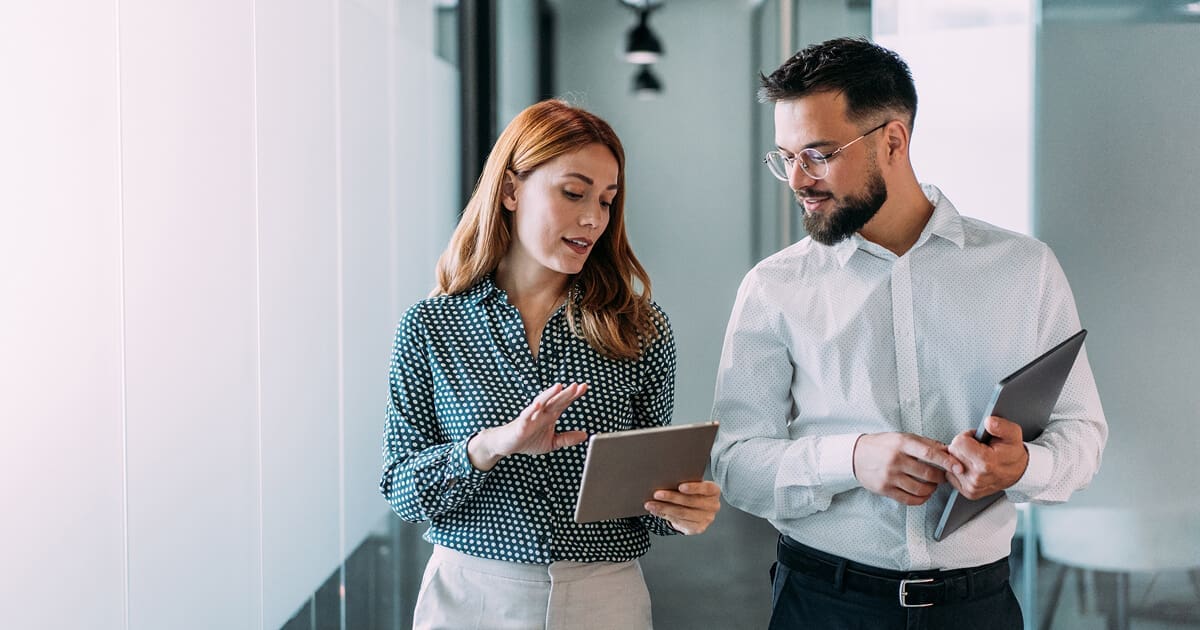 Two business professionals walking through an office hallway while discussing information on a tablet