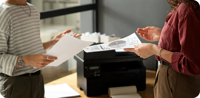 Employees reviewing printed reports beside a network printer to monitor print usage and control printing costs.