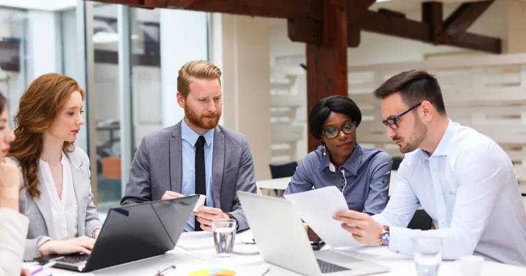 Group of professionals collaborating in a modern office meeting, reviewing documents and working on laptops around a conference table.