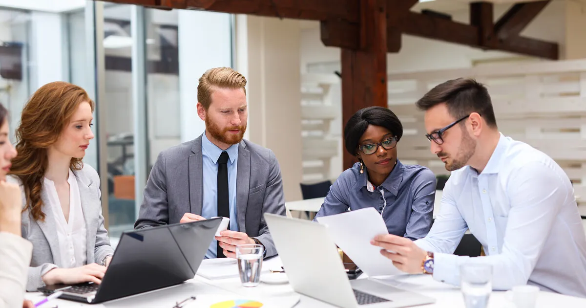 Group of professionals collaborating in a modern office meeting, reviewing documents and working on laptops around a conference table.